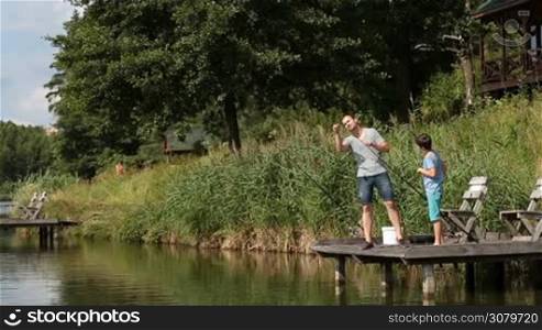 Caring father untangling hook with bait from fishing line of son&acute;s rod while fishing together on wooden pier on freshwater pond over beautiful summer landscape background. Dad and teenage boy getting ready for angling on lake.