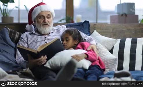 Caring grandfather with grey beard and in santa hat reading a book to his sleepy cute mixed race granddaugther as they lie in bed on christmas eve. Affectionate grandpa cuddling his adorable girl while reading bedtime story at home on Xmas.