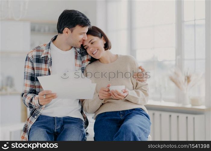 Caring husband embraces wife with love, have glad expressions, holds paper documents, discuss banking procedures, study bills, drink tea or coffee, pose indoor, enjoy calm domestic atmosphere