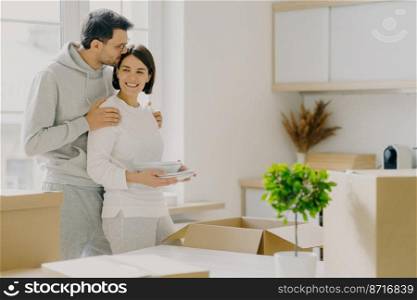 Caring male embraces his wife with love and tenderness, kisses in head, pose together in kitchen, move into new rented flat, unpack carton boxes with plates, enjoy relocation. Moving Day concept