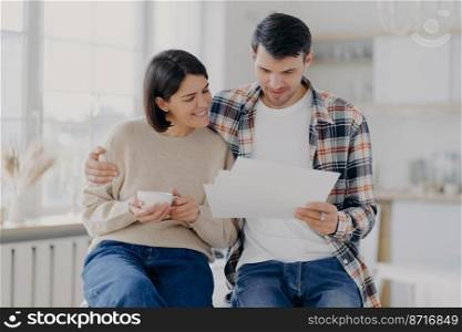 Caring man embraces his wife, look through documents with happy expressions, study payment bills, drinks coffee, dressed in casual wear, pose in spacious room with blurred background. Family and work