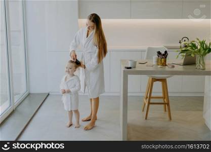 Caring mother combs hair of her small daughter wear white bathrobes pose against kitchen interior stand bare feet at floor. Beautiful little curly girl after taking shower. Mom making hairstyle to kid