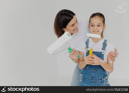 Caring mother embraces small daughter with love, paint walls of new house together, hold paint rollers, have happy faces, isolated over white background with empty space. Home makeover, renovation