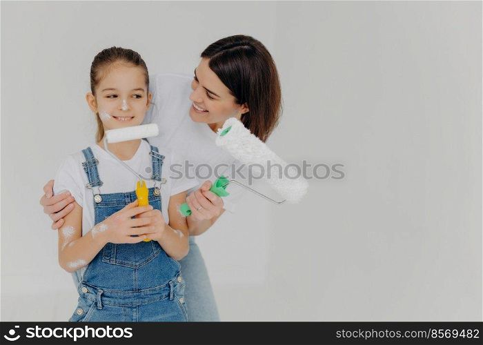 Caring mother embraces small daughter with love, paint walls of new house together, hold paint rollers, have happy faces, isolated over white background with empty space. Home makeover, renovation