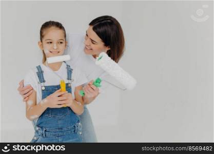 Caring mother embraces small daughter with love, paint walls of new house together, hold paint rollers, have happy faces, isolated over white background with empty space. Home makeover, renovation