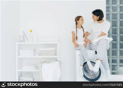 Caring mother talks with daughter, pose on washing machine, surrounded with white linen, do washing together, being in laundry room at home. Happy adult housewife thanks adorable girl for help