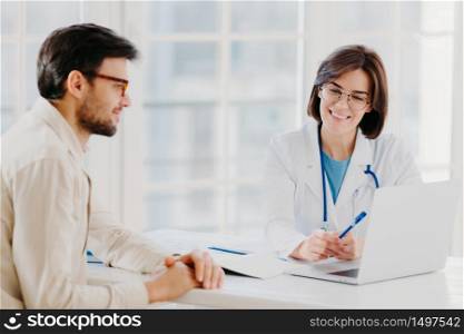 Caring woman doctor gives prescriptions to patient, sit together at table and look at laptop computer, fill up application form online. Sick man visit healthcare clinic, gets medical consultancy