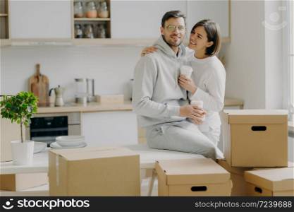 Caring woman embraces husband with love, drink takeaway coffee, pose in modern kitchen with unpacked boxes around, move into new apartment for living, rent flat, unpack belongings, have break
