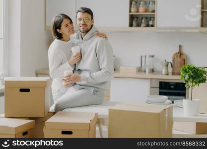 Caring woman embraces husband with love, drink takeaway coffee, pose in modern kitchen with unpacked boxes around, move into new apartment for living, rent flat, unpack belongings, have break