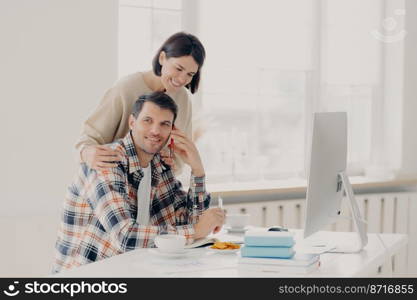 Caring woman helps her husband entrepreneur who has telephone conversation, tries to soleve working issues, writes information in notepad and works on computer. Family couple manage finances