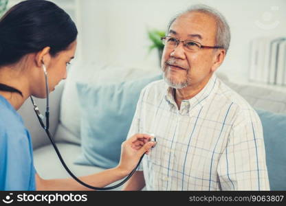 Caring young female doctor examining her contented senior patient with stethoscope in living room. Medical service for elderly, elderly sickness, declining health.. Caring young female doctor examining her contented senior patient.