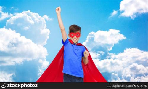 carnival, childhood, power, gesture and people concept - happy boy in red super hero cape and mask showing fists over blue sky and clouds background
