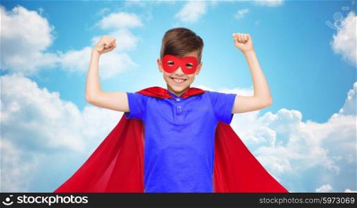 carnival, childhood, power, gesture and people concept - happy boy in red super hero cape and mask showing fists over blue sky and clouds background