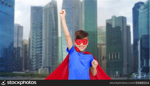 carnival, childhood, power, gesture and people concept - happy boy in red super hero cape and mask showing fists over city background