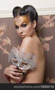 carnival portrait of stunning brunette woman with creative hairdo and glossy golden make-up, posing with naked shoulders and baroque sparkle mask in the hand
