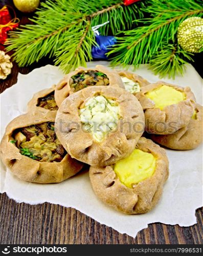 Carols of rye flour filled with cheese, potatoes and mushrooms on paper, Christmas balls, fir branches on the background of dark wooden board
