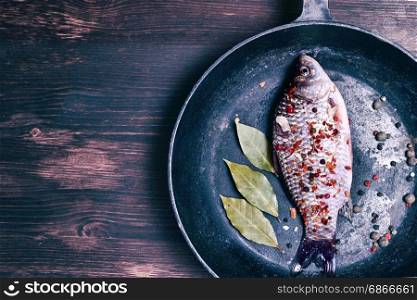 Carp fish in a black cast-iron frying pan on a brown wooden background, empty space on the left