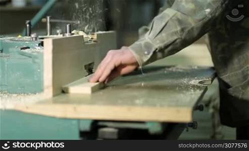 Carpenter&acute;s hands working on electric cutter in workshop, close up on the component part. Experinced joiner decoring wood plank on electric bench cutter and putting down finished product on stack of wooden planks. Selective focus
