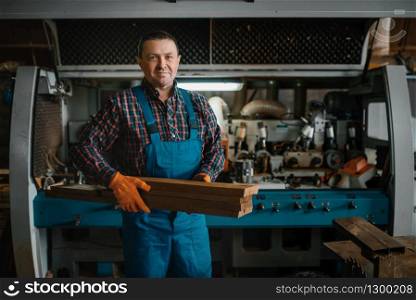 Carpenter in uniform holds boards, woodworking machine on background, lumber industry, carpentry. Wood processing on factory, forest sawing in lumberyard