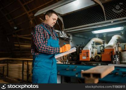 Carpenter in uniform holds notebook, woodworking machine on background, lumber industry, carpentry. Wood processing on factory, forest sawing in lumberyard