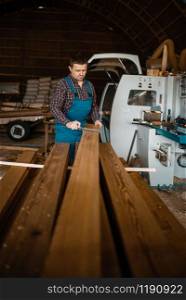 Carpenter in uniform measures the board with caliper, woodworking machine on background, lumber industry, carpentry. Wood processing on factory, forest sawing in lumberyard. Carpenter in uniform measures board with caliper