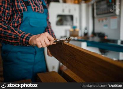 Carpenter in uniform measures the board with caliper, woodworking machine on background, lumber industry, carpentry. Wood processing on factory, forest sawing in lumberyard