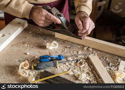 Carpenter man scraping curled wood scraps with hand plane tool and wooden plank.