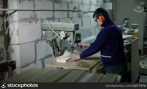 Carpenter wearing protective goggles and earmuffs boring holes in wooden board using vertical drilling machine. Drilling piece of wood with electric milling cutter in carpentry.