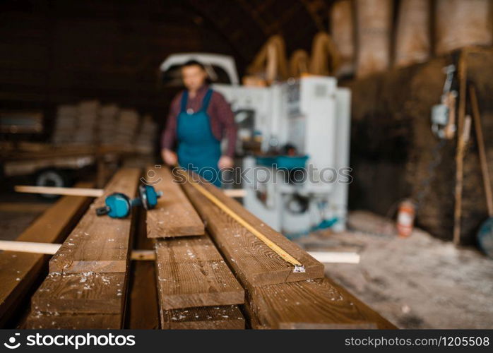 Carpenter with measuring tape measures boards, woodworking machine on background, lumber industry, carpentry. Wood processing on factory, forest sawing in lumberyard