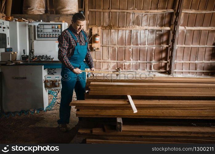 Carpenter with measuring tape measures boards, woodworking machine on background, lumber industry, carpentry. Wood processing on factory, forest sawing in lumberyard. Carpenter with measuring tape measures boards