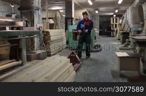 Carpenter working on manual forklift pallet staked with wooden boards in workshop. Joiner pushing fork pallet truck with stack of planed planks at carpentry shop.