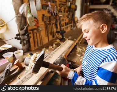carpentry, woodwork, building and people concept - happy little boy with hammer hammering wood plank at workshop