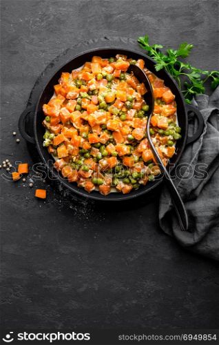 Carrot braised with fresh green peas in creamy milk sauce in stewpan, vegetable saute on black background, top view