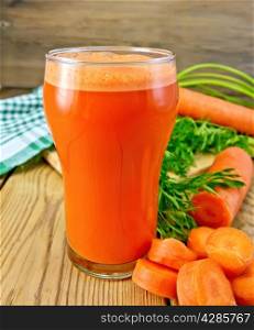 Carrot juice in a tall glass, carrots with greens, a napkin on the background of wooden boards