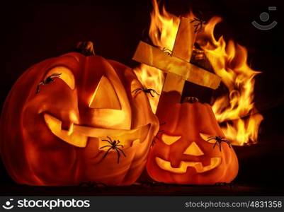 Carved glowing pumpkin with creepy spiders, bright burning fire near cross, terrible decoration for Halloween holiday party, horror and magic concept