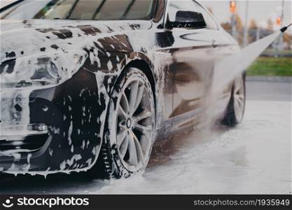 Carwashing. Worker spraying water from high pressure washer on black luxury car covered in white soap foam, cleaning vehicle with shampoo at self-service car wash station outdoors. Process of professional car wash with chemical detergent and high pressure washer