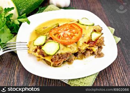 Casserole of minced meat, tomato and zucchini in a plate on a napkin, fork, garlic on a background of a dark wooden board