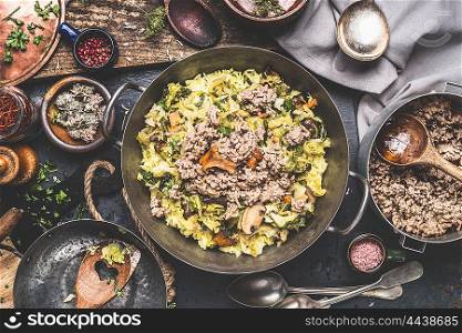 Casserole with rice , chopped cabbage and minced meat on rustic kitchen table with wooden spoons and bowls with flavoring and spices, top view.