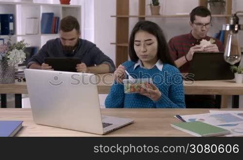 Casual asian brunette businesswoman eating healthy business lunch in office interior as her colleagues working on background. Freelancer woman eating fresh vegetables mix at workplace while working.