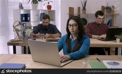 Casual asian businesswoman stretching arms while sitting at desk in front of laptop in office. Relaxed female freelancer stretching her arms in busy office with male business partners on background.