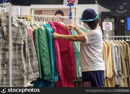 Casual Asian woman wears beanie with protective mask choosing jacket clothes on clothing rack in fashion store at shopping mall