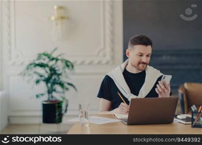 Casual businessman multitasks with smartphone, notes important contacts, and watches business webinar on social media. Freelancer works on laptop from home office.