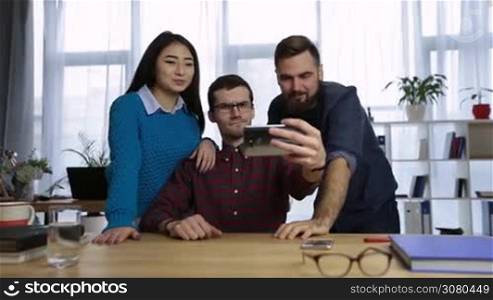 Casual diverse business team taking self-portrait with smart phone in startup office. Smiling business people making funny facial expressions while taking selfie on mobile phone in office. Dolly.