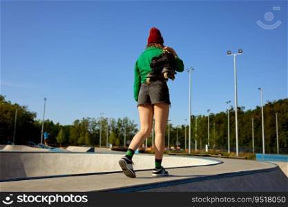 Casual girl carrying rollerblades in hands walking to training class. Outdoor rollerdrome for practicing freestyle speed riding. Casual girl carrying rollerblades in hands walking to training class