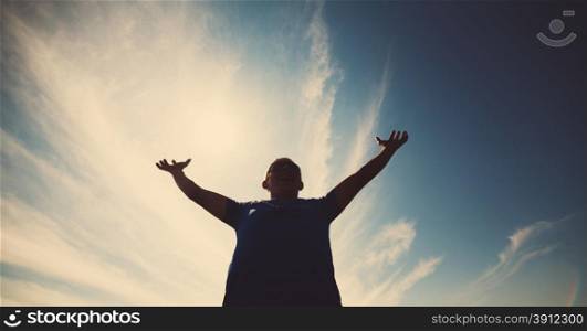 Casual man looking very happy with his arms up against blue sky in backlight . Young casual man gesturing success