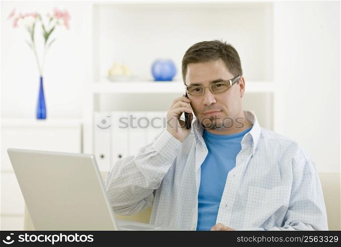 Casual man using laptop computer at home and calling on phone.