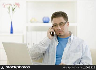 Casual man using laptop computer at home and calling on phone.