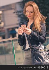 Casual modern young woman checking her social media, using smartphone outdoor. Fashionable female wearing jeans jacket with phone.. Fashionable woman using phone outdoor
