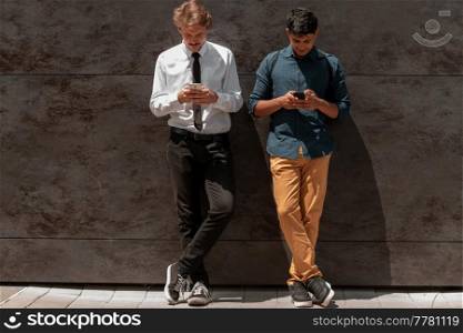 Casual multiethnic startup business men one of them is Indian using smartphone during a break from work in front of the grey wall outside. High-quality photo. Casual multiethnic startup business men one of them is Indian using smartphone during break from work in front of grey wall outside