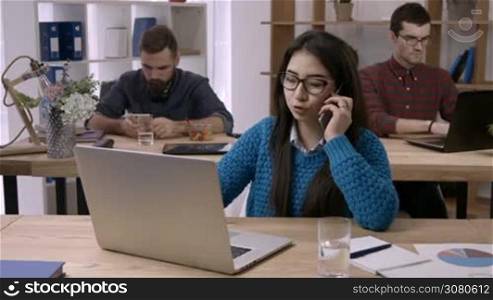 Casual multitasking working day of independent businesswoman. Beautiful young asian woman in eyeglasses talking on mobile phone and using laptop while sitting at desk at her workplace in modern office